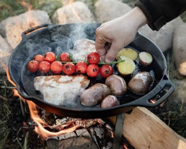 Tigaie din fonta, pe suport, deasupra focului cu bucati de carne, cartosi si rosii cherry