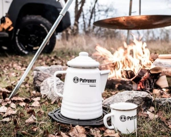 Percolator si cana Petromax albe asezate pe iarba langa foc